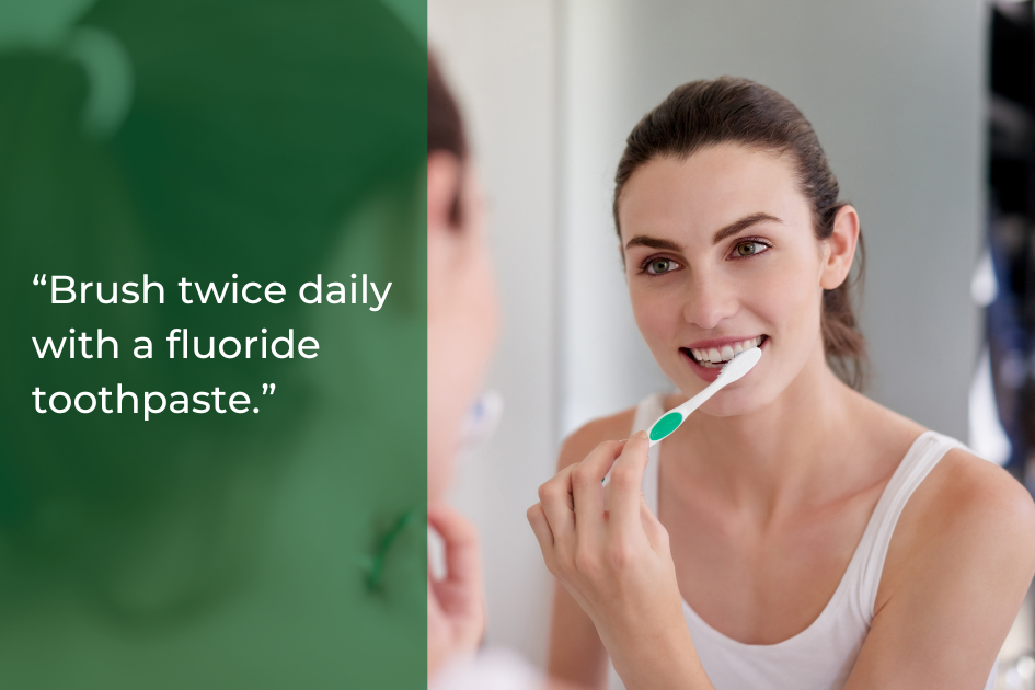 A woman in Taylor TX brushes her teeth with a white and green toothbrush in front of a mirror. Text on the image reads: “Brush twice daily with a fluoride toothpaste for better oral health.”.