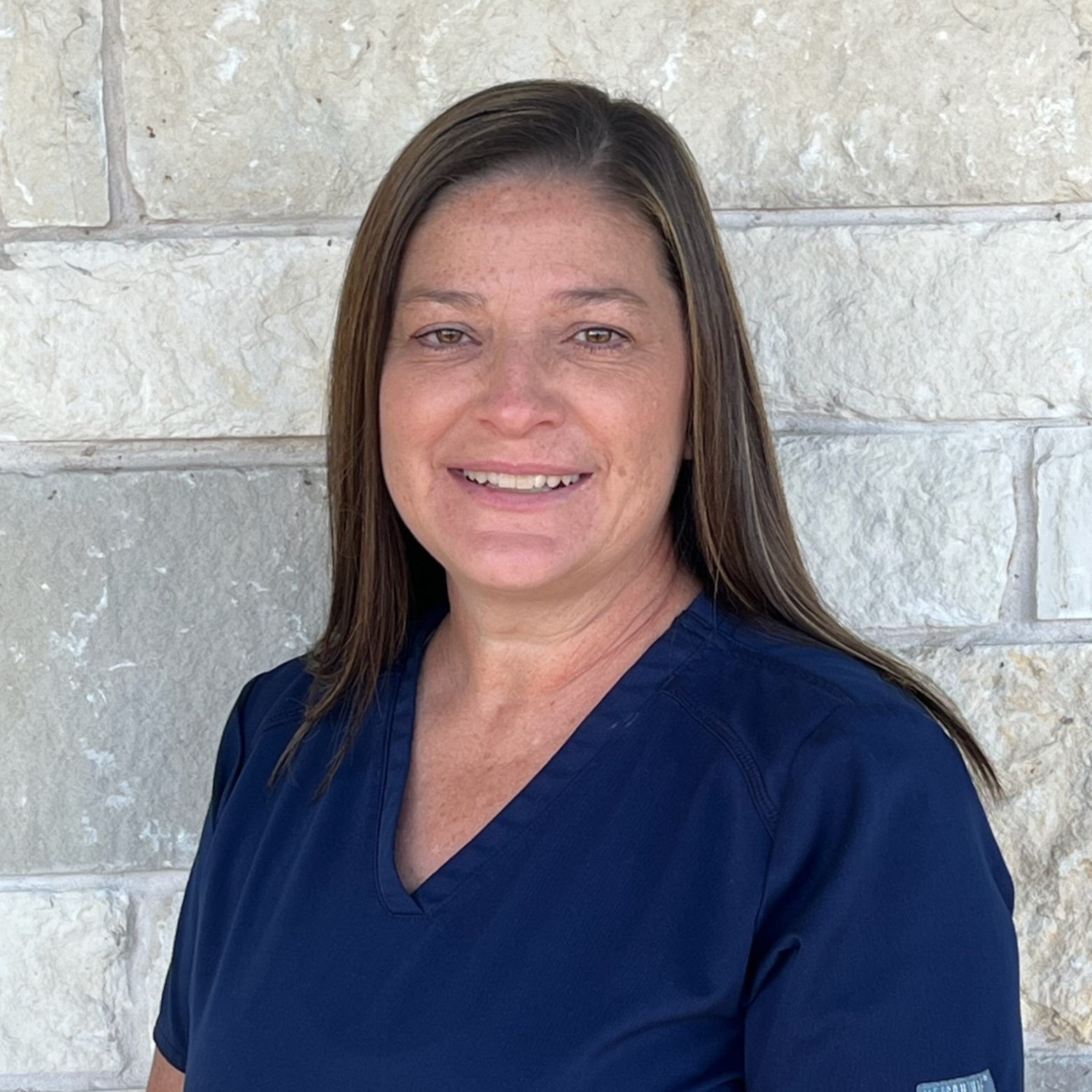A woman with straight brown hair wearing navy blue scrubs stands in front of a light-colored stone wall, smiling at the camera—meet the team member who makes a difference every day.
