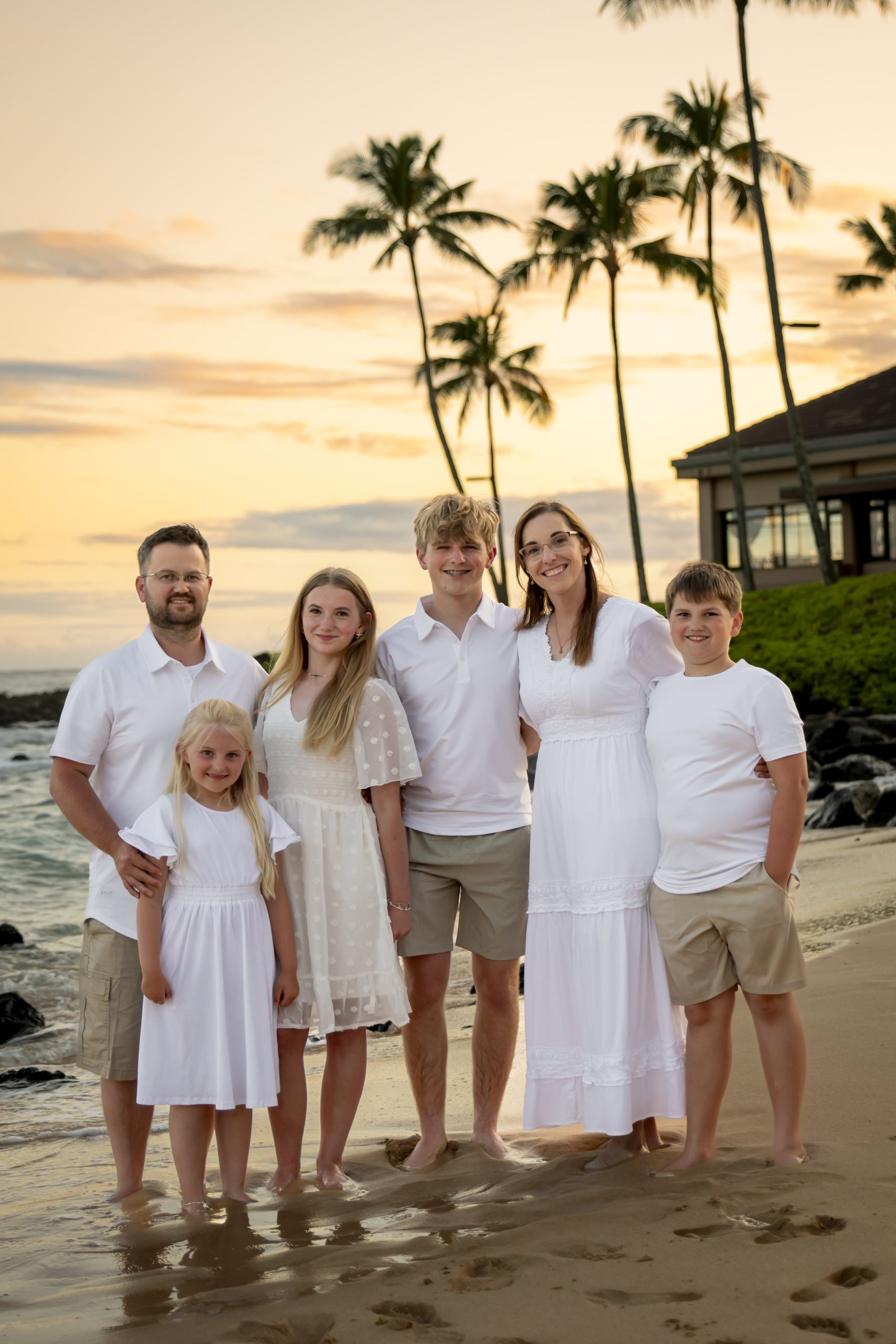 A family of six poses together on a beach at sunset, all wearing white clothing. Palm trees and a building are visible in the background, perfect for those searching "Dentist Near Me" while enjoying a relaxing coastal getaway.
