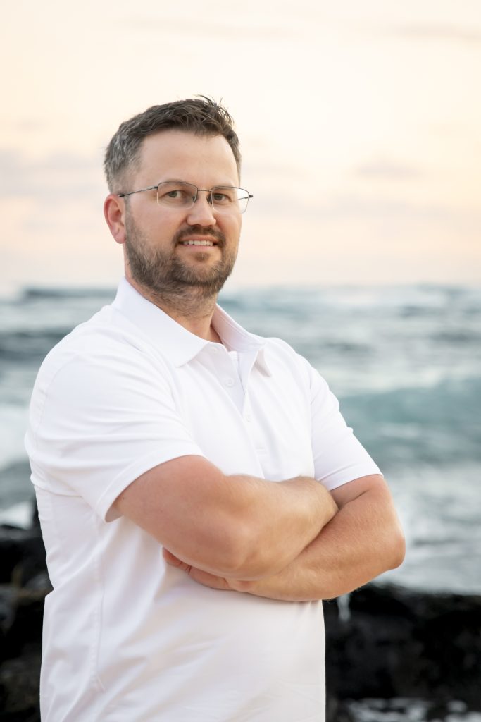 A man with glasses and a beard stands with arms crossed, wearing a white polo shirt, in front of an ocean sunset—looking confident and approachable, like a trusted Dentist Near Me.