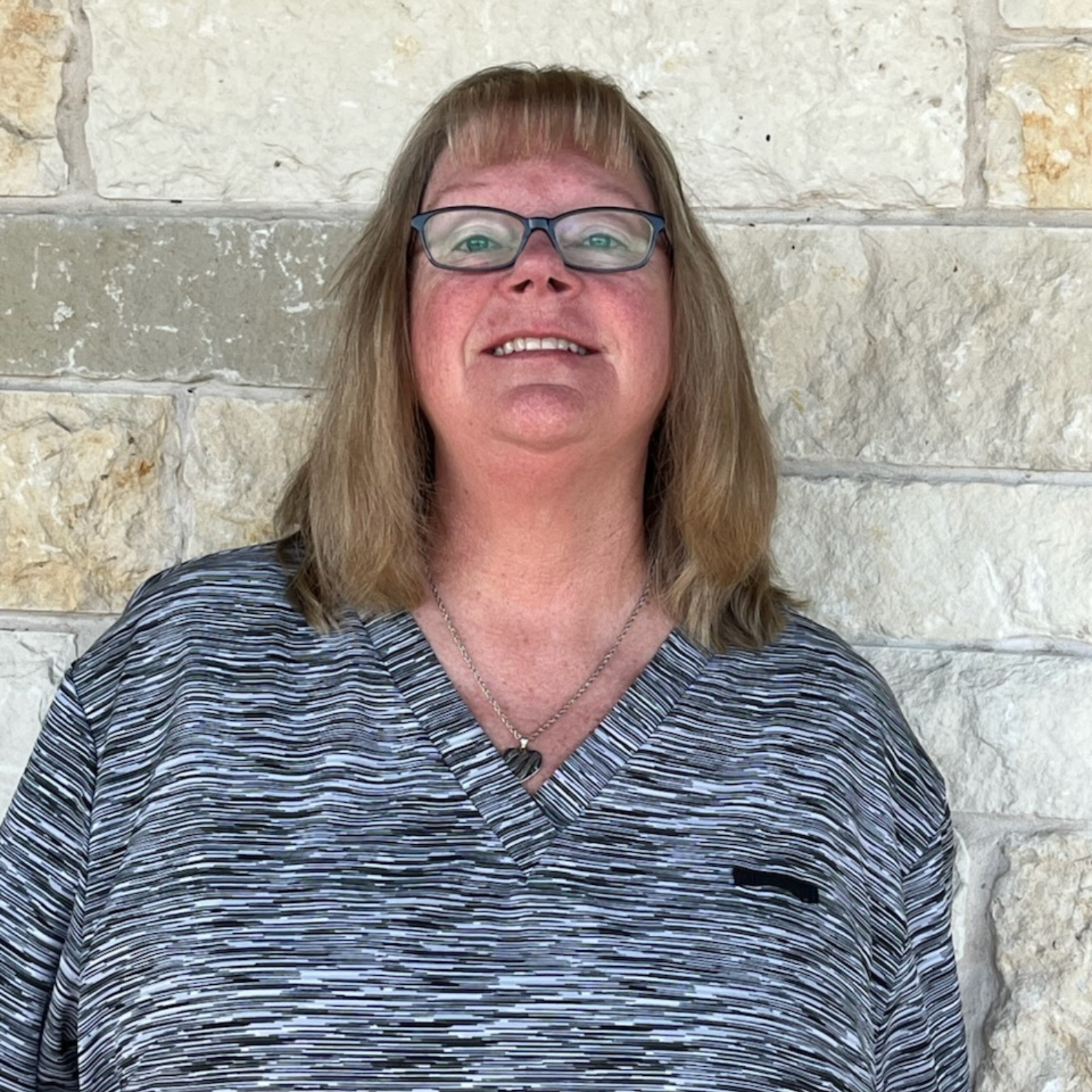 Meet the Team: A woman with shoulder-length light brown hair and glasses stands in front of a stone wall, smiling. She is wearing a striped gray and black top and a necklace.